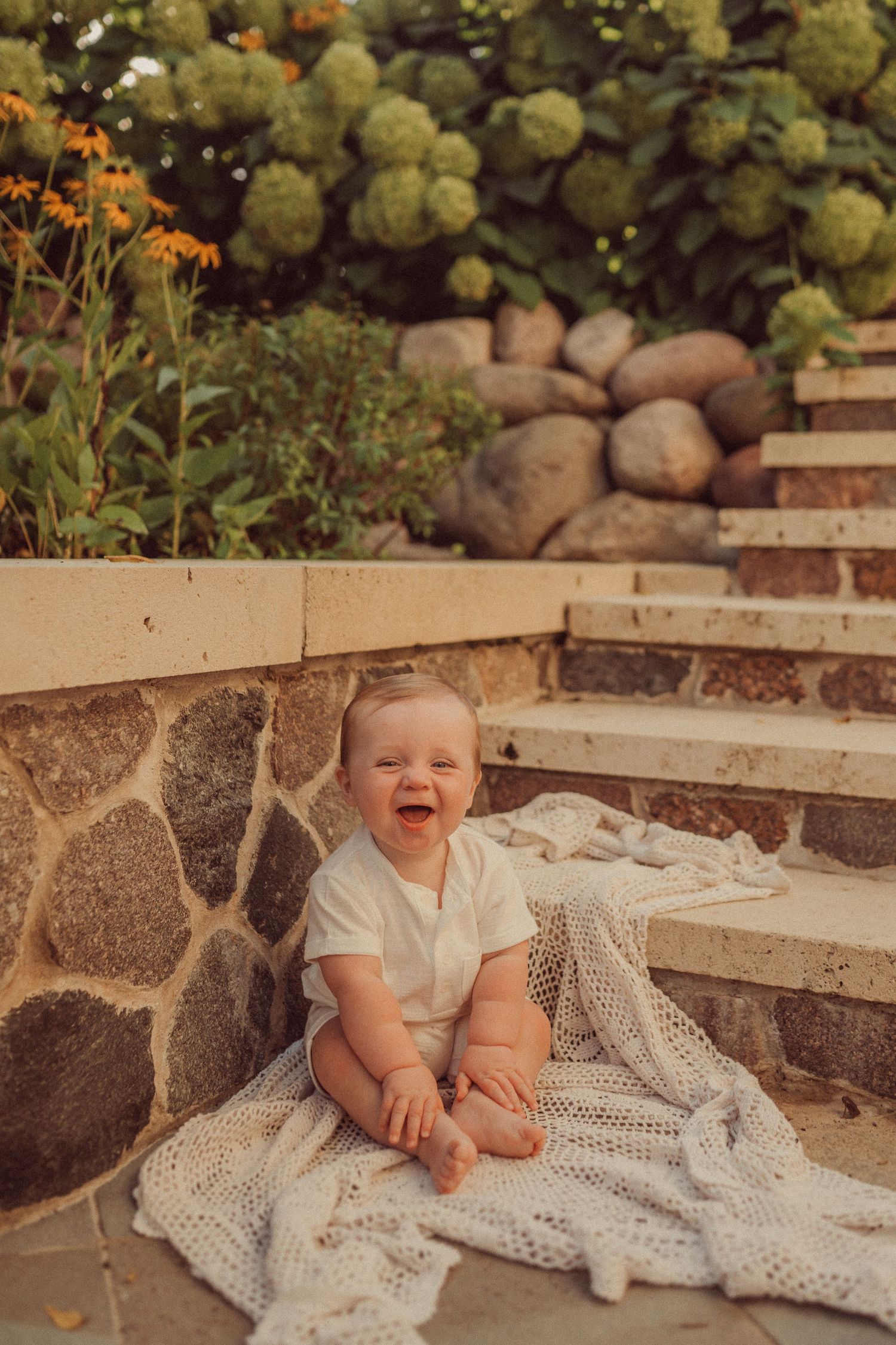 Happy baby sits on lace blanket on stone steps with hydrangeas and flowers in background.