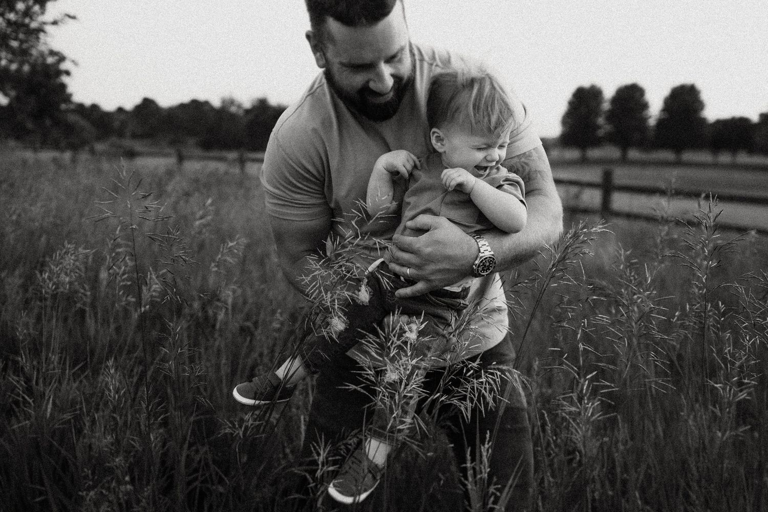 Black and white intimate moment between family members in tall grass with rustic fencing behind.