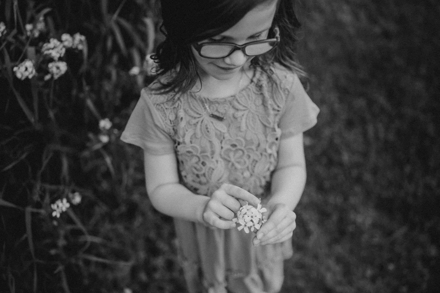 Black and white portrait of a child looking down while standing among garden flowers.
