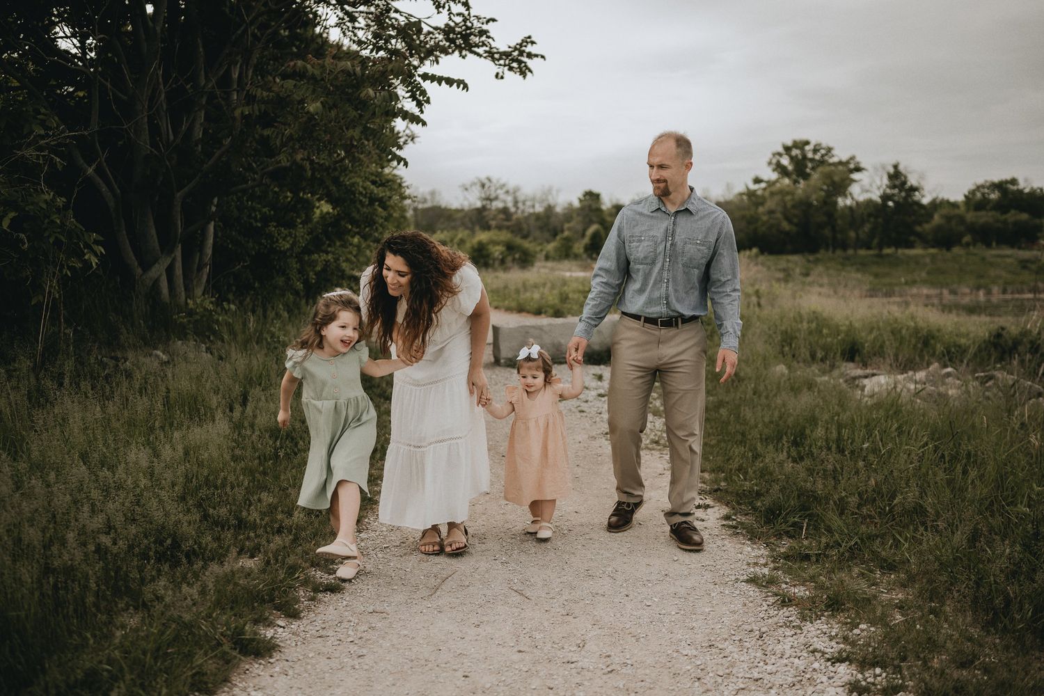 A carefree family strolls together down a winding dirt path through tall grasses on a cloudy summer day.