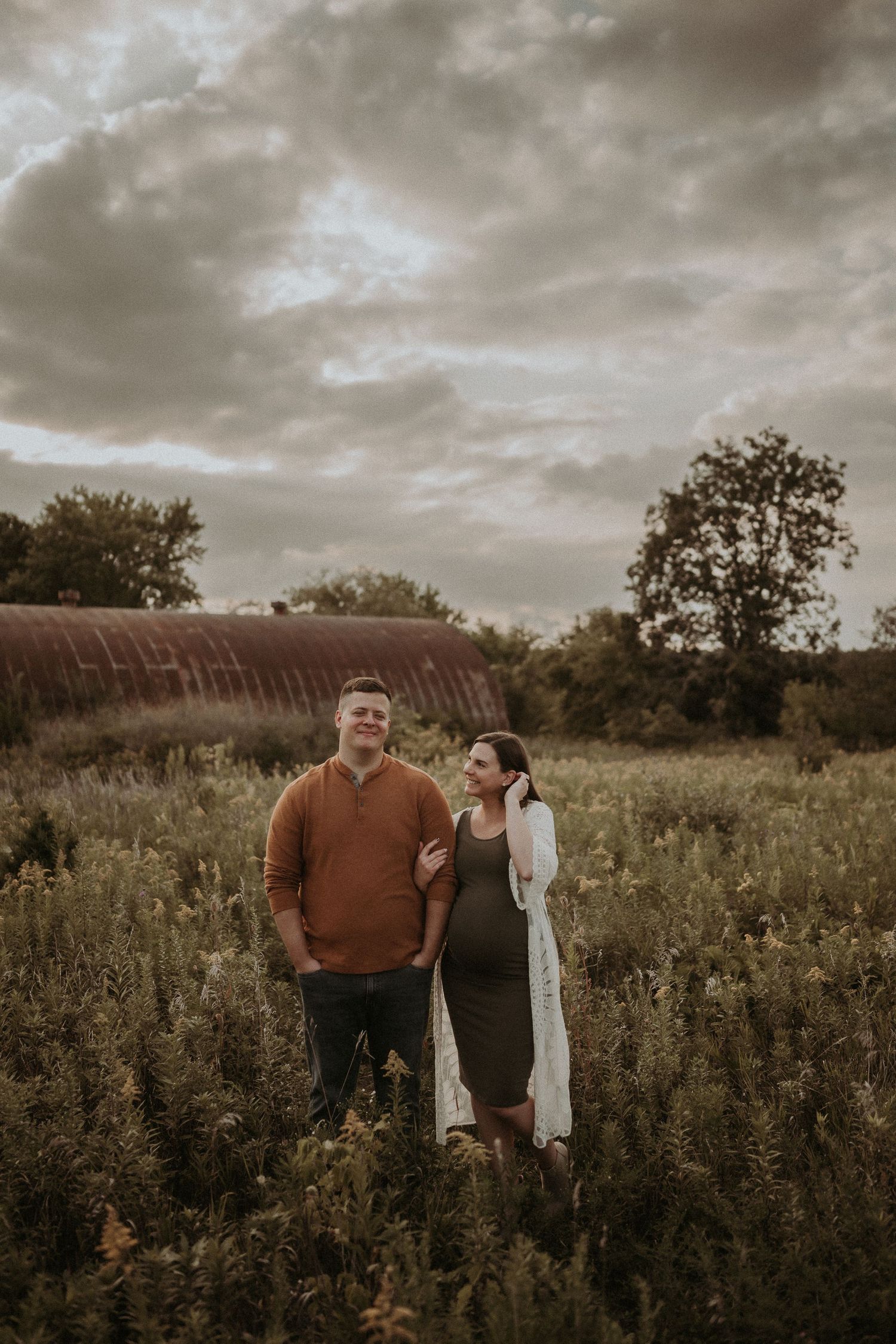 Couple poses together in autumn field with rustic barn and dramatic cloudy sky.