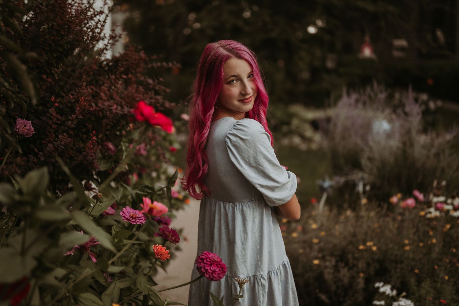 A person in a flowing dress poses in a garden with vibrant pink flowers and red hair.