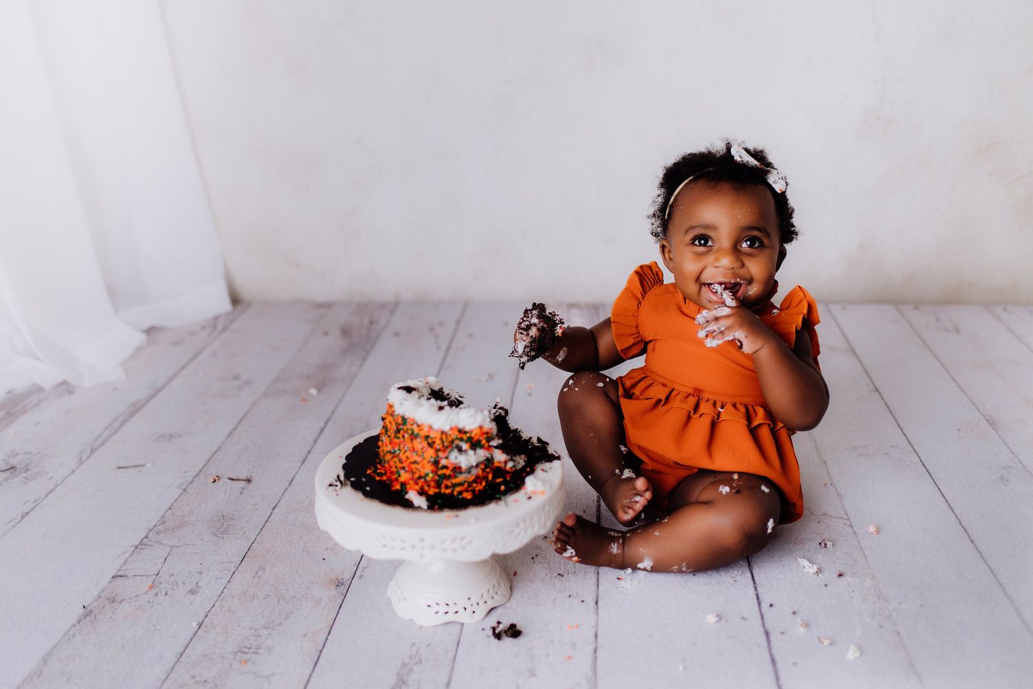 Small child in orange dress enjoying a messy cake smash photoshoot on white wooden floor.