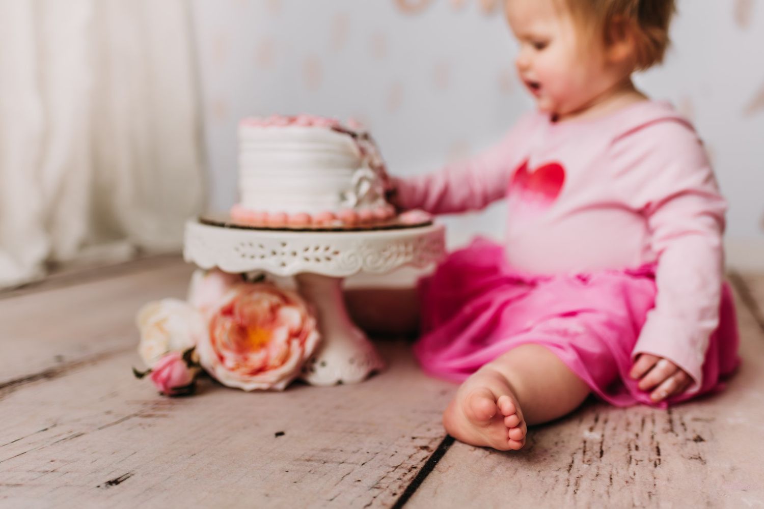 Pink-dressed figure sits beside decorated cake on rustic wooden floor with floral accents.