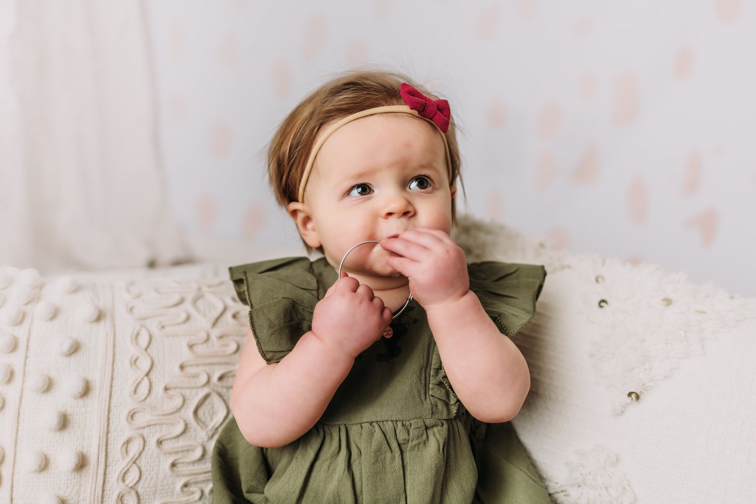 Baby in olive dress with red bow touches face while sitting on cream pillows.