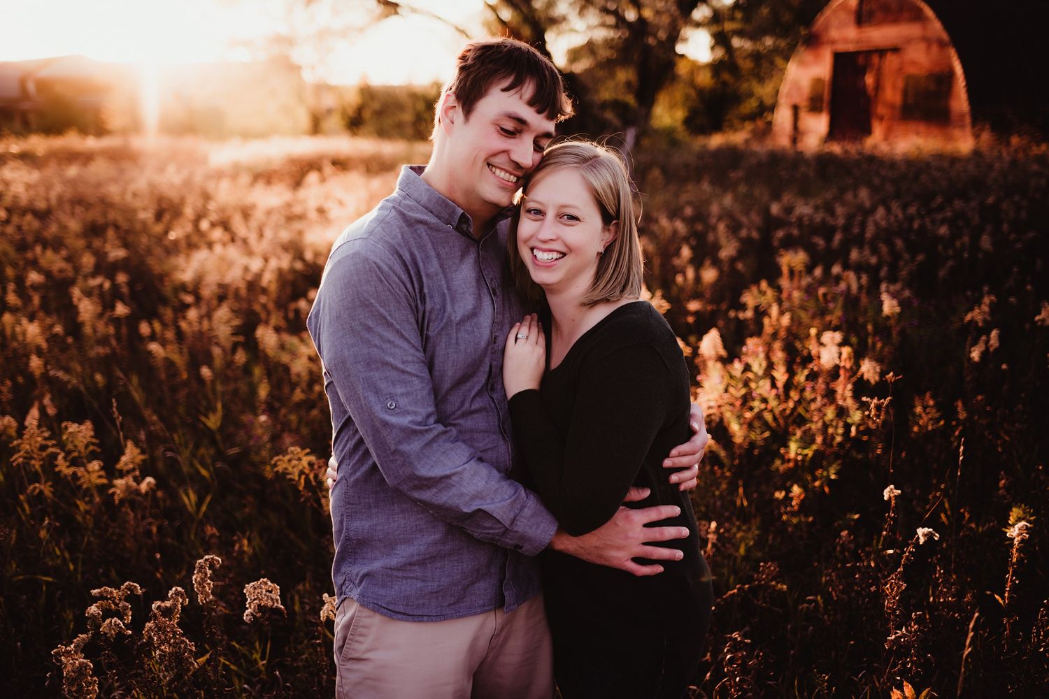 A couple shares an intimate moment in a field at sunset with a red barn in the background.