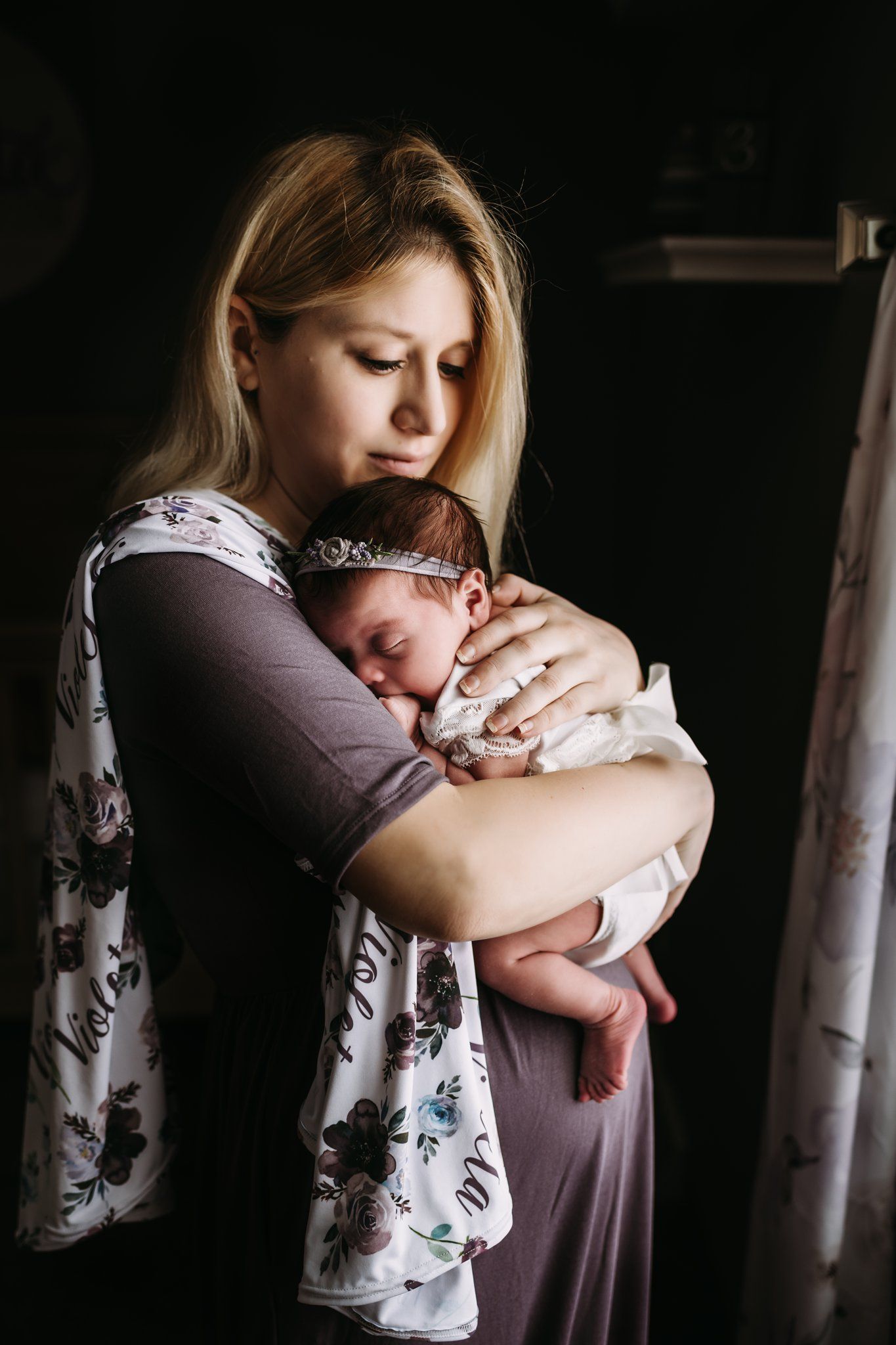 Mother in floral dress cradles sleeping newborn in dark room.
