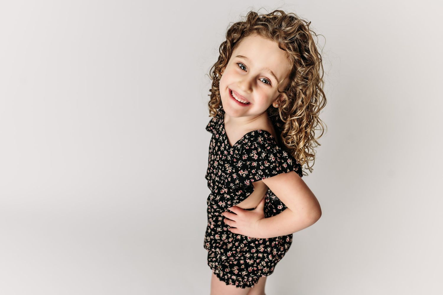 A child strikes a fun pose in a floral romper against white backdrop.