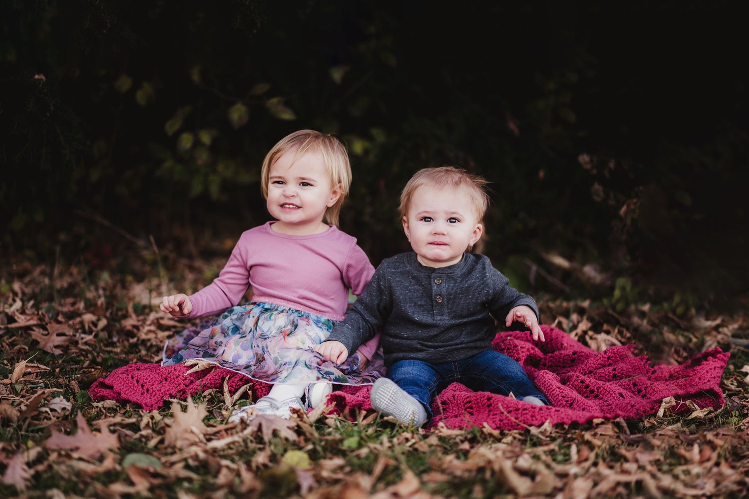 Two toddlers sit together on red blanket surrounded by fallen leaves.
