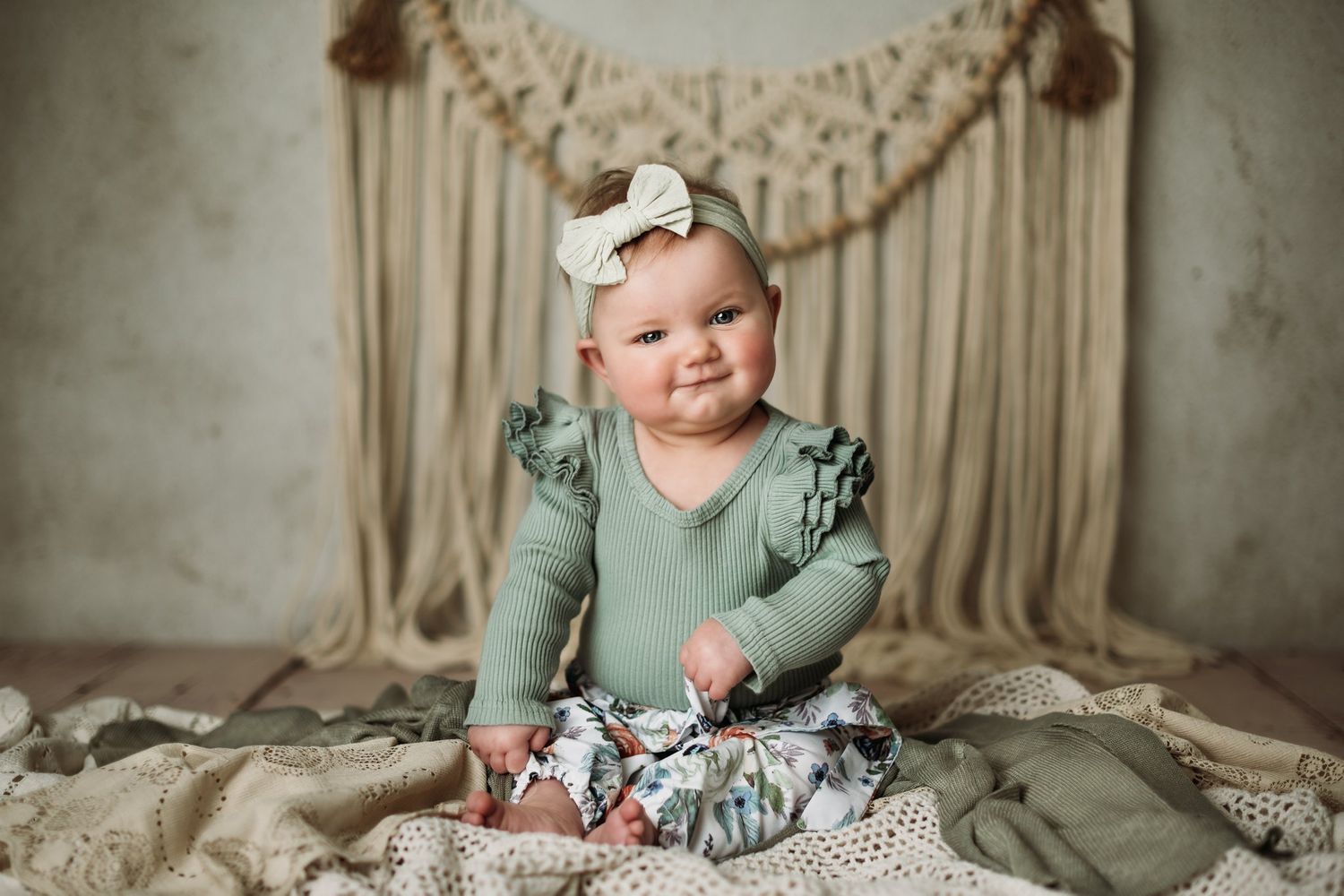 Baby wearing sage green outfit smiles while sitting on lace blanket with macrame wall hanging.