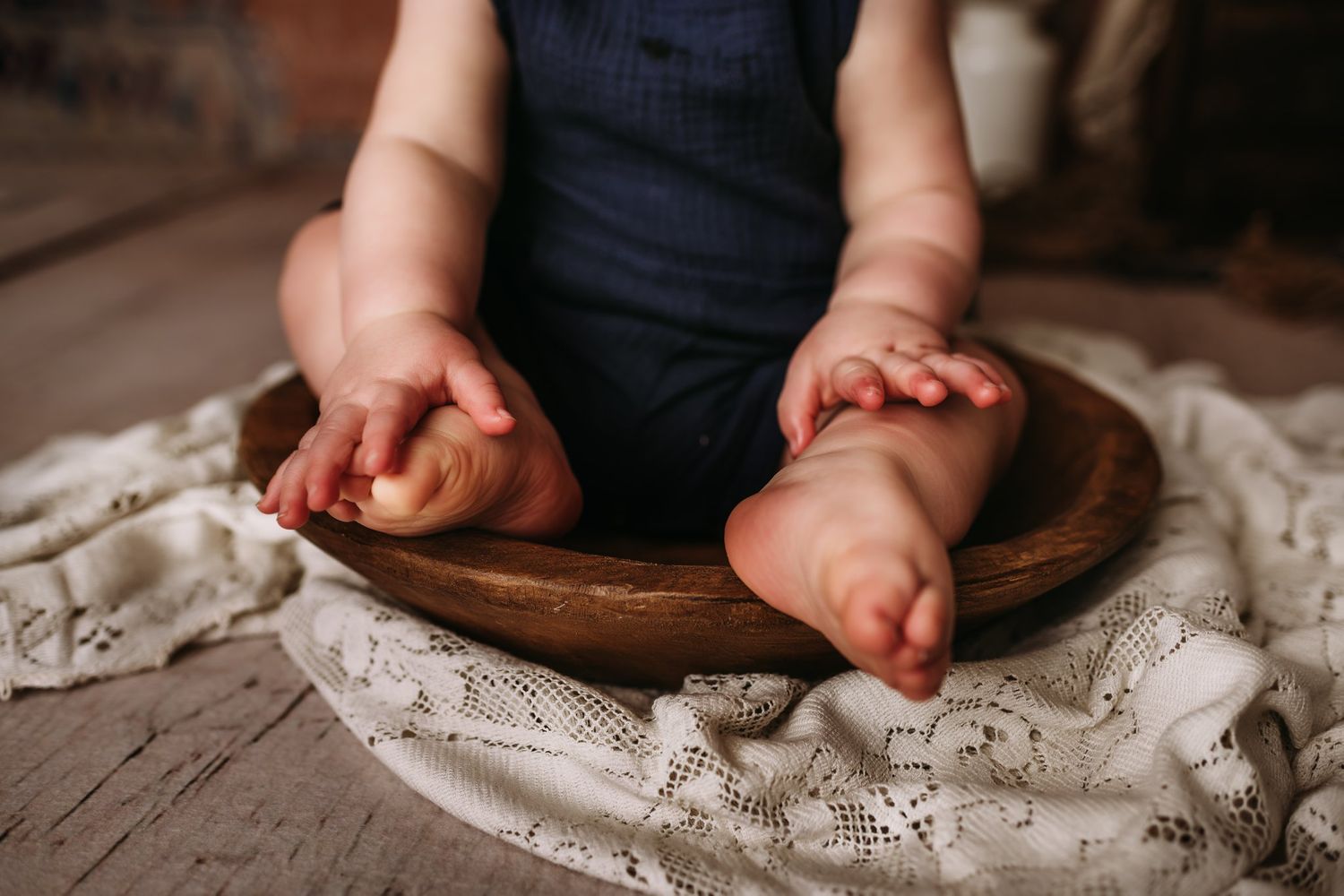 Close-up of small feet and hands in wooden bowl on white lace fabric against wooden floor.