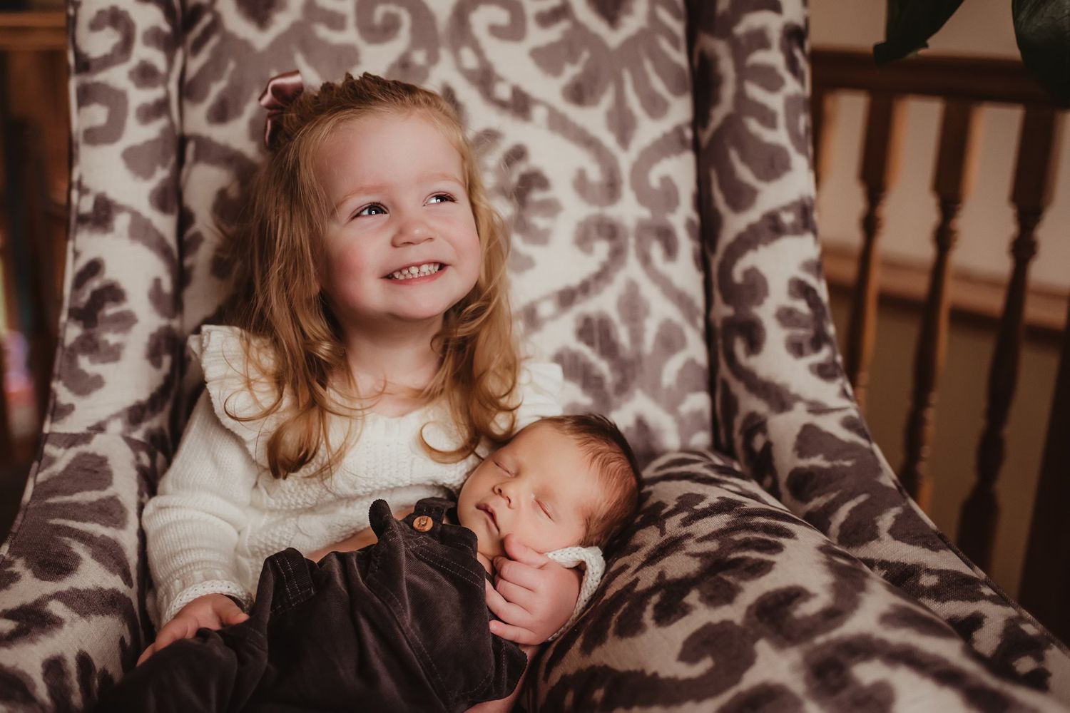 Smiling young child sitting in a patterned armchair while carefully holding a sleeping newborn.