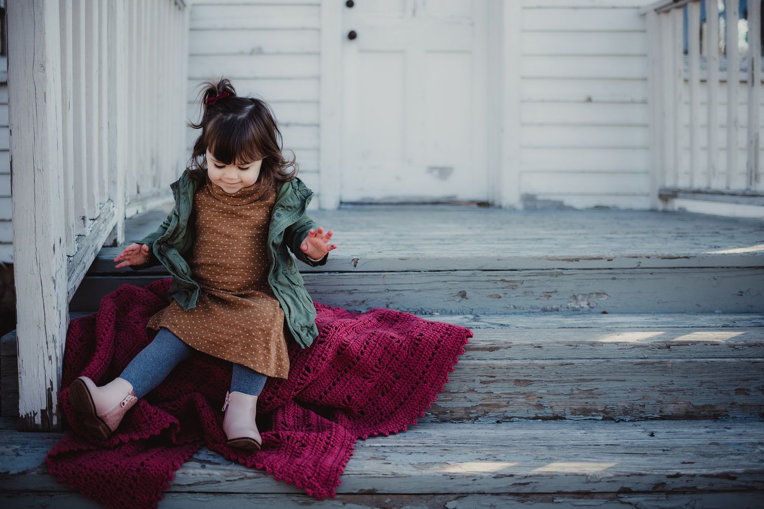 A child relaxes on porch steps with a burgundy blanket wearing autumn clothing.