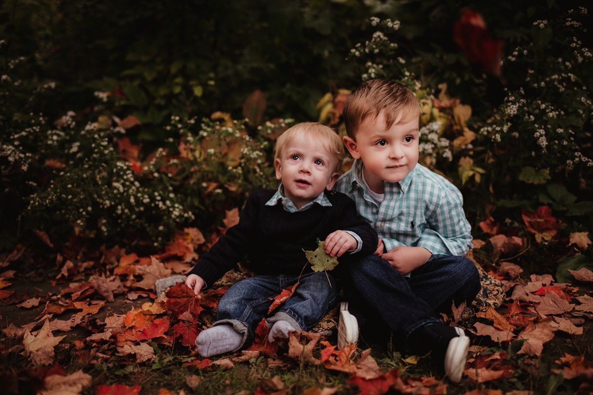 Two young children sitting together on ground covered with red autumn leaves.