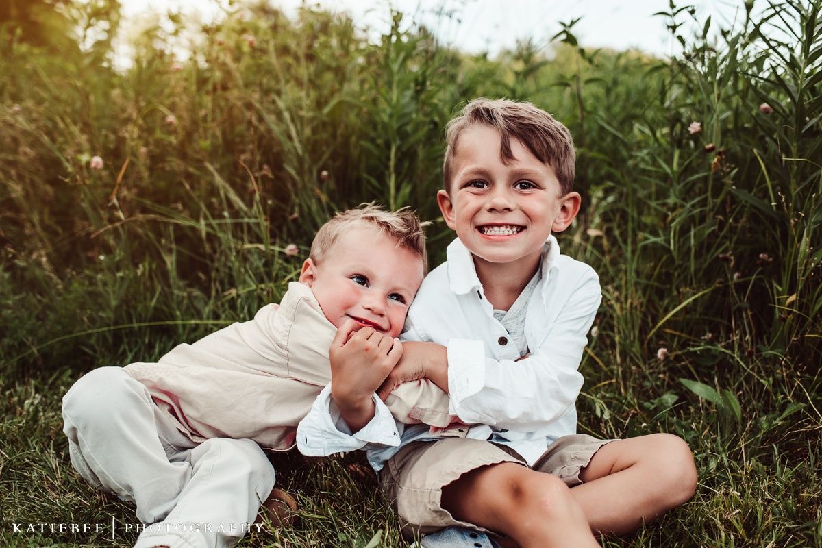 Two children sitting together and laughing in tall grass during golden hour.
