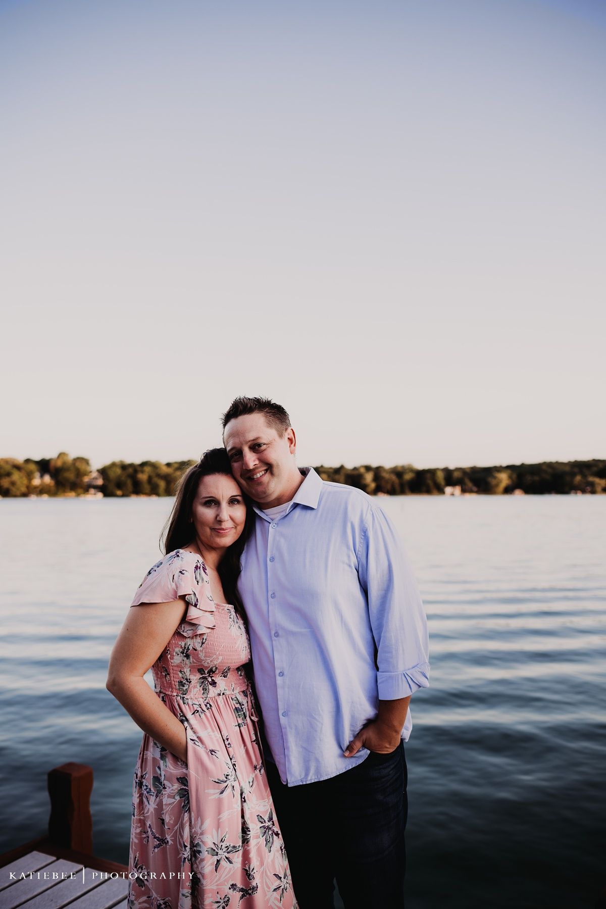 Couple poses together at sunset by a lake with a rocky shoreline in the background.