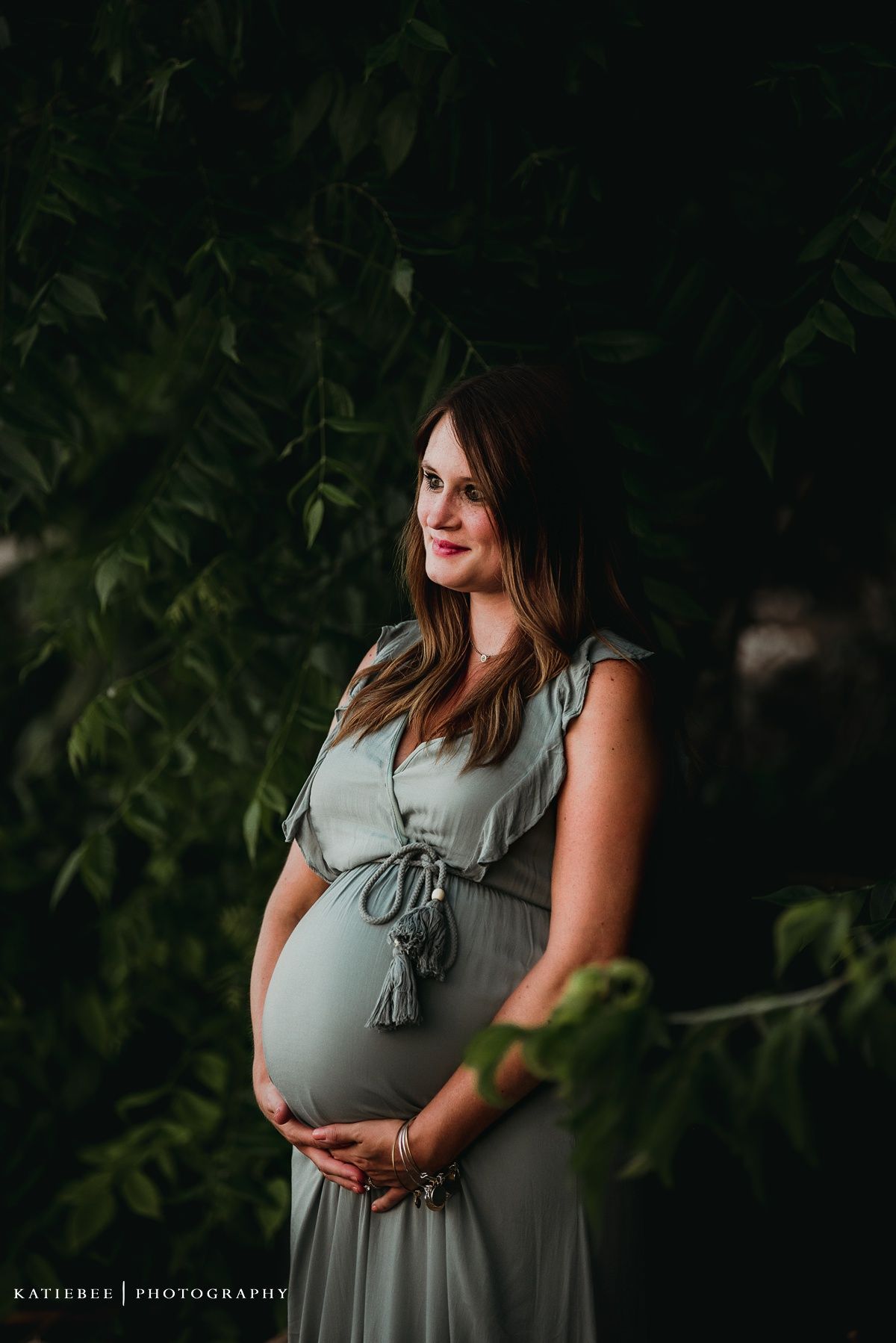 An expectant mother in a sage green dress poses against a leafy backdrop.