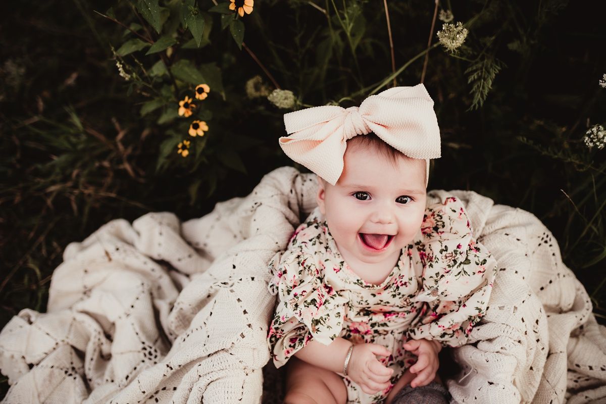 Joyful baby wearing floral outfit and bow headband laughs while laying on blanket outdoors.