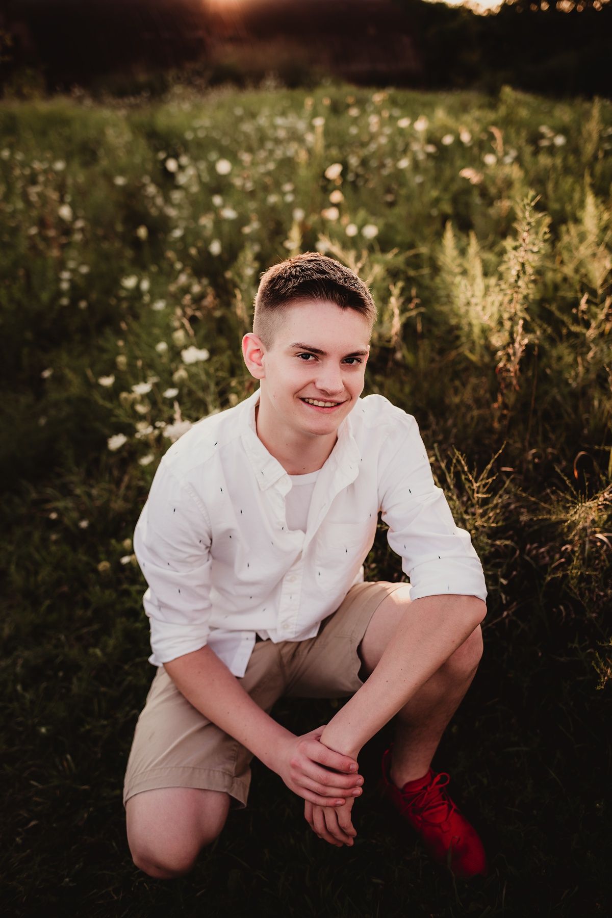 A person in white shirt and khaki shorts crouching in a wildflower field at sunset.