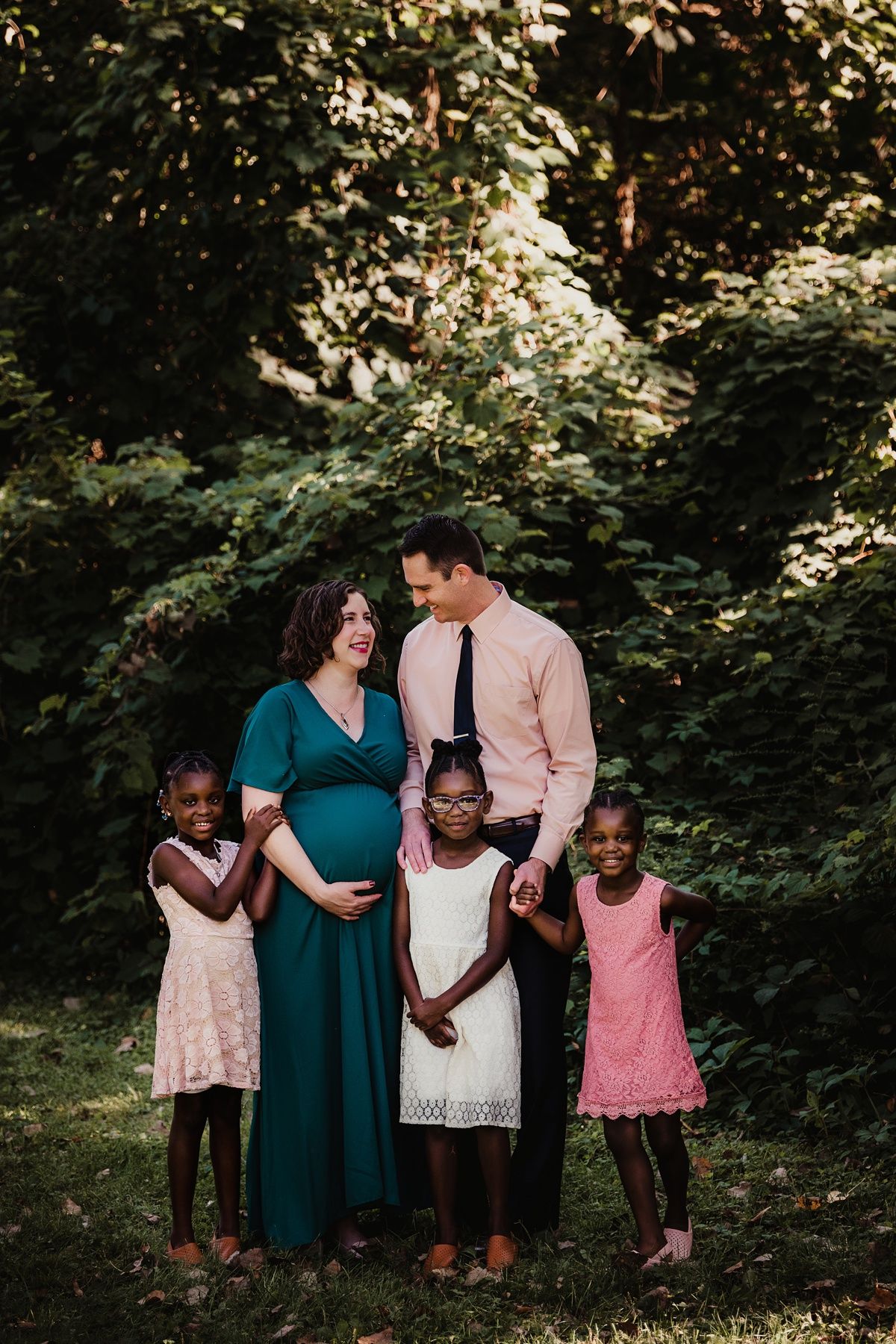A blended family poses together outdoors, featuring a couple in teal and pink with three children in lace dresses.