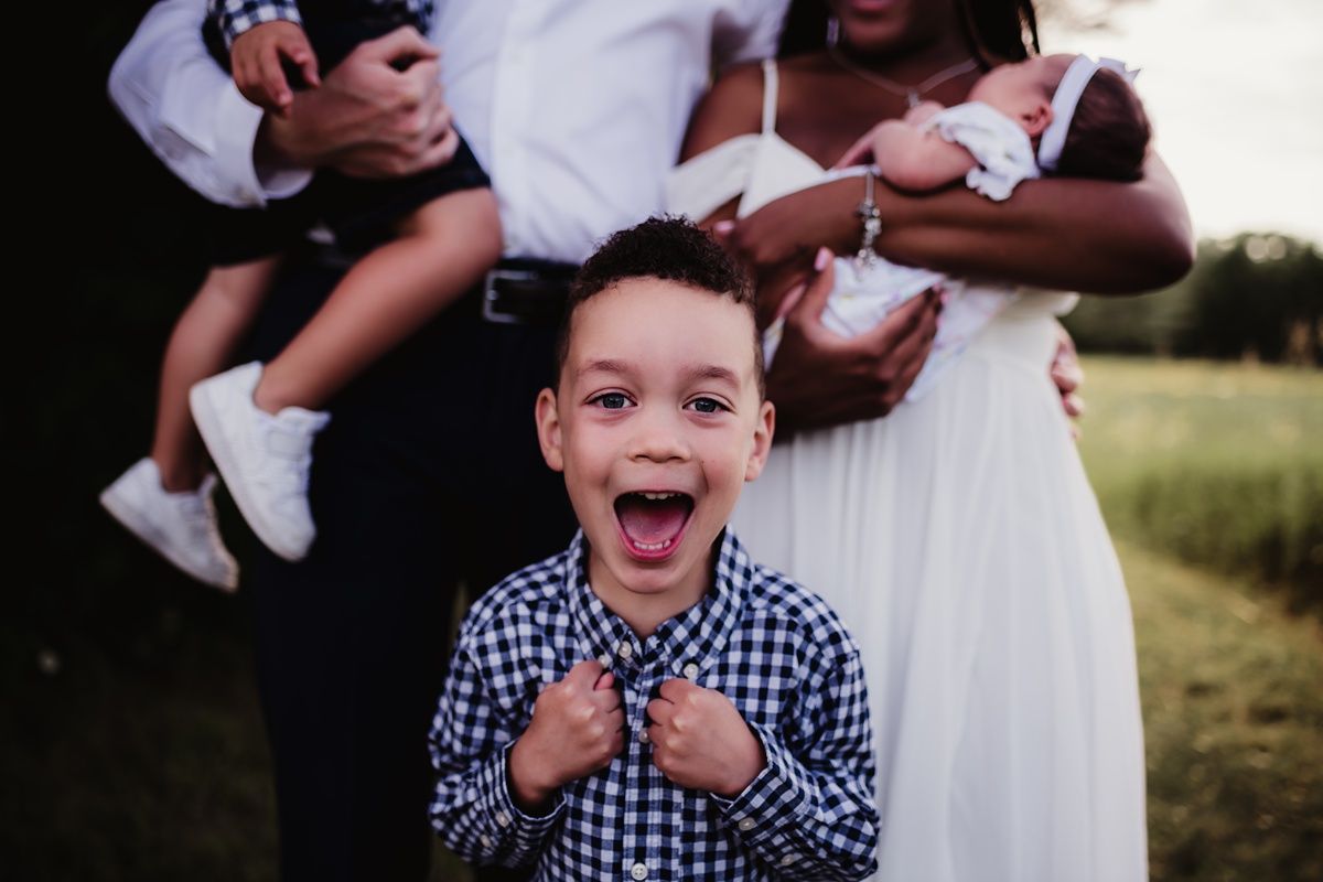 Joyful outdoor family photo with excited child in foreground and others holding baby in background.