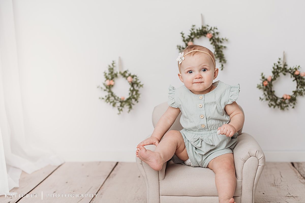 Baby sitting on wooden floor touching face with decorative wreaths in background.
