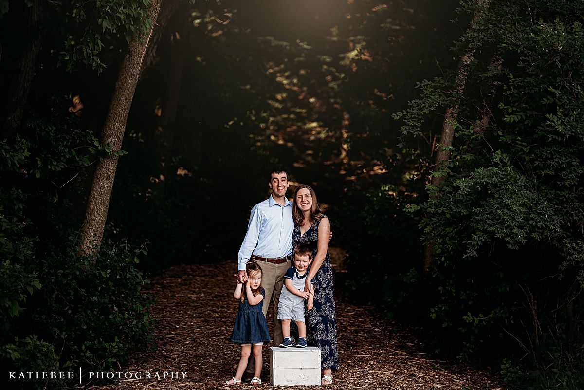 A family stands together on a wooden box for a portrait in a wooded setting.
