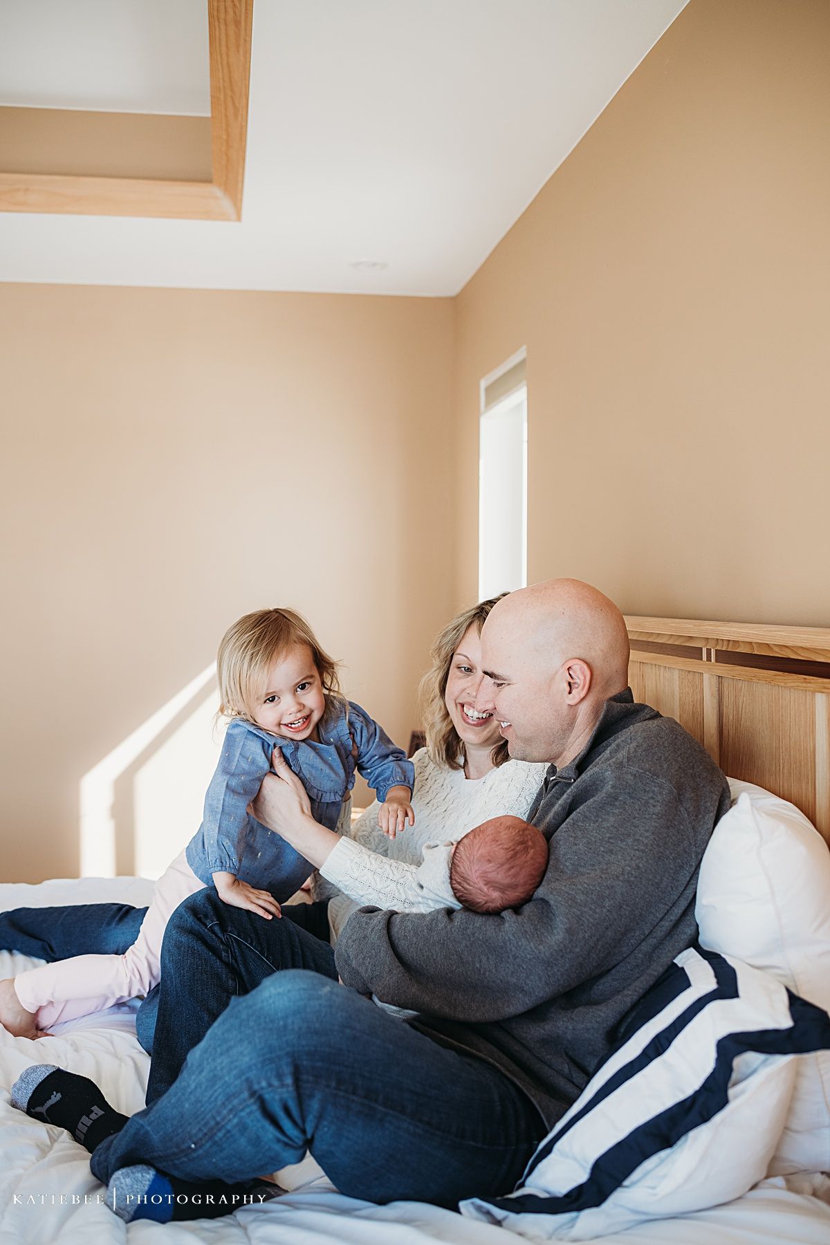A family sits together on a bed in a sunlit room with wooden furniture.