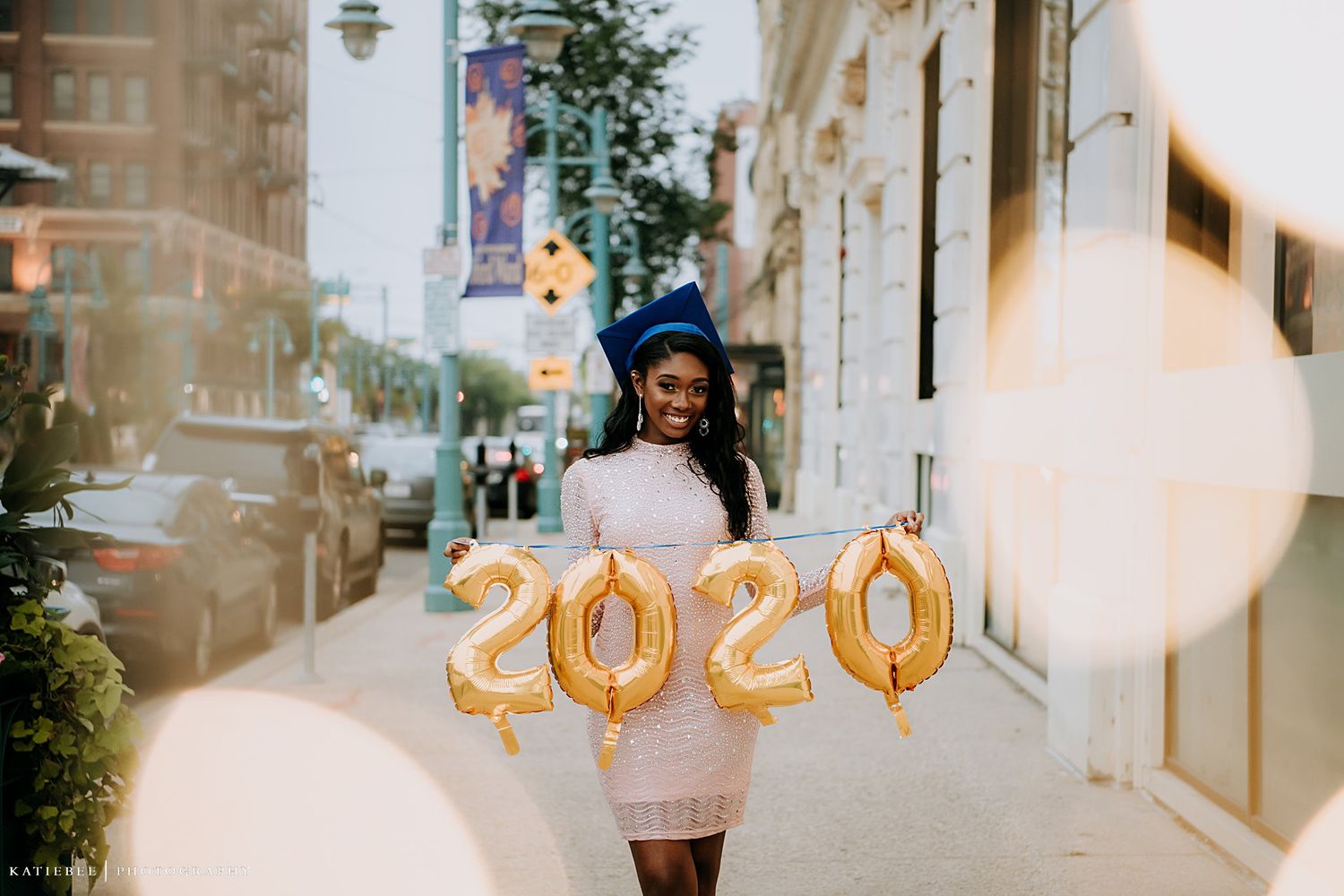 Graduate posing on city street with gold 2020 number balloons and evening lighting.