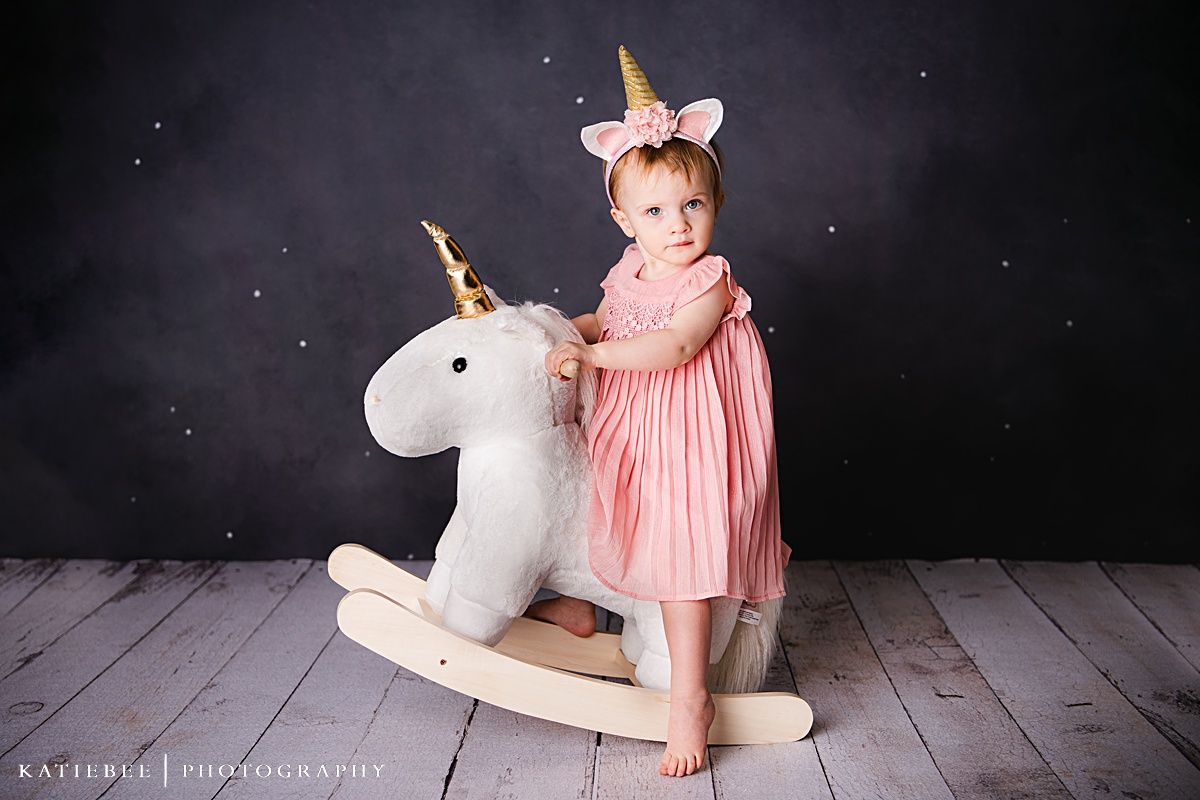 A smiling toddler in a pink dress plays on a white rocking unicorn toy.