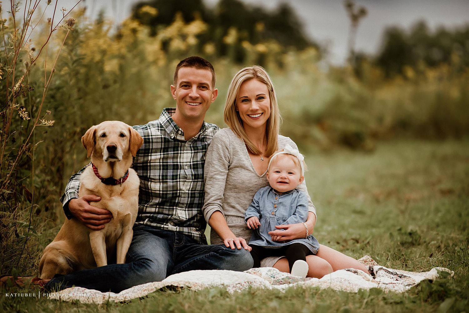 Family portrait outdoors with their golden retriever dog sitting on blanket in golden field.