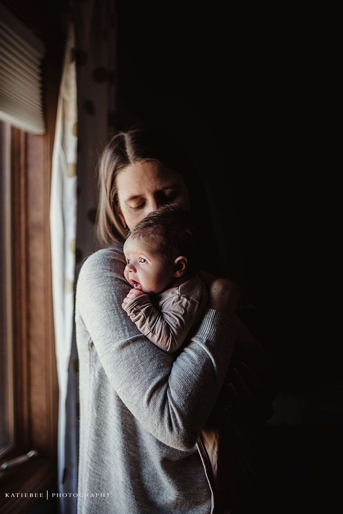 Moody window light portrait of caregiver embracing infant in grey toned setting.
