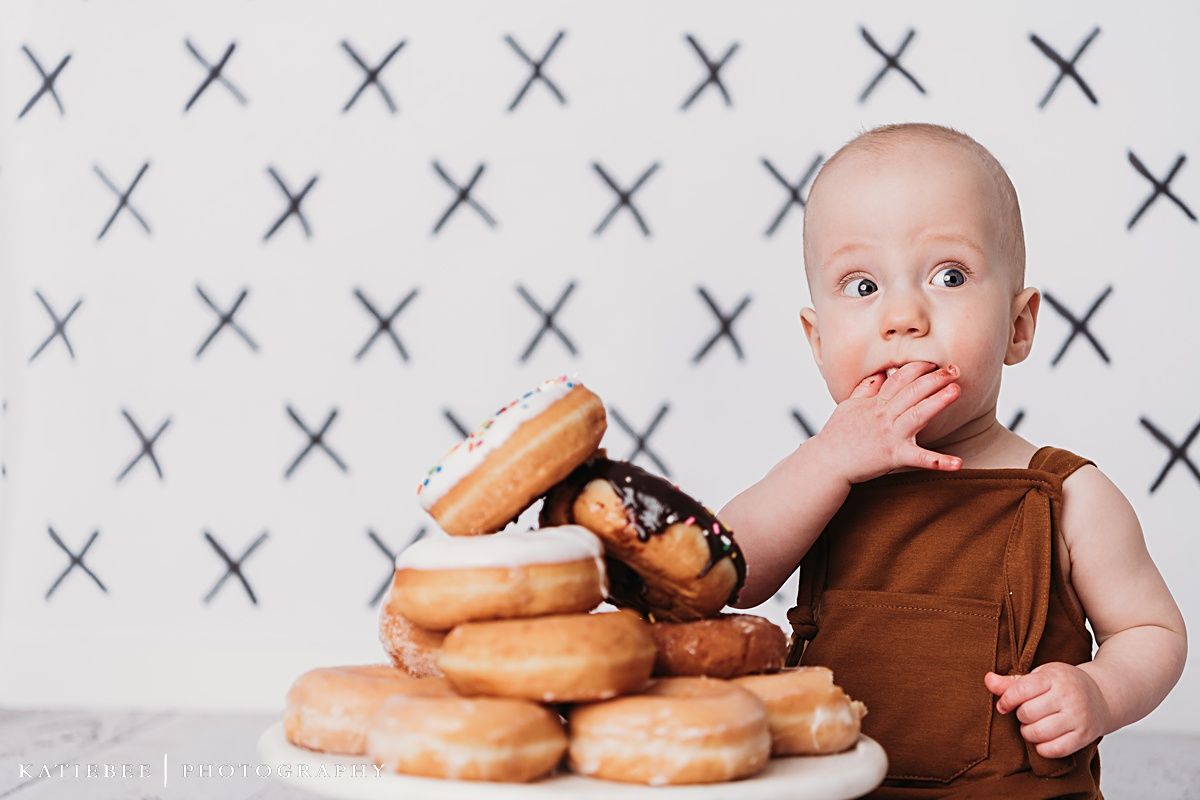 Baby in brown outfit reaches for stack of donuts on cake stand against X-pattern background.