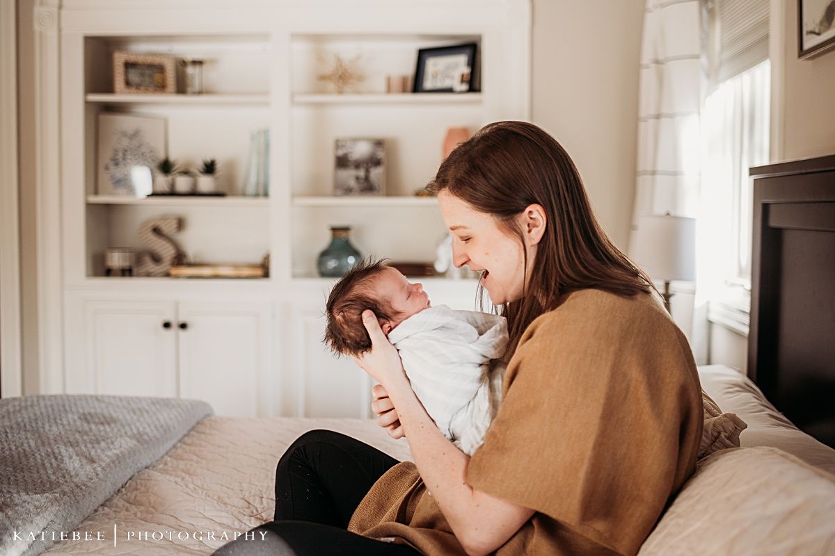 Parent and infant share tender moment while sitting on bed near built-in shelving.