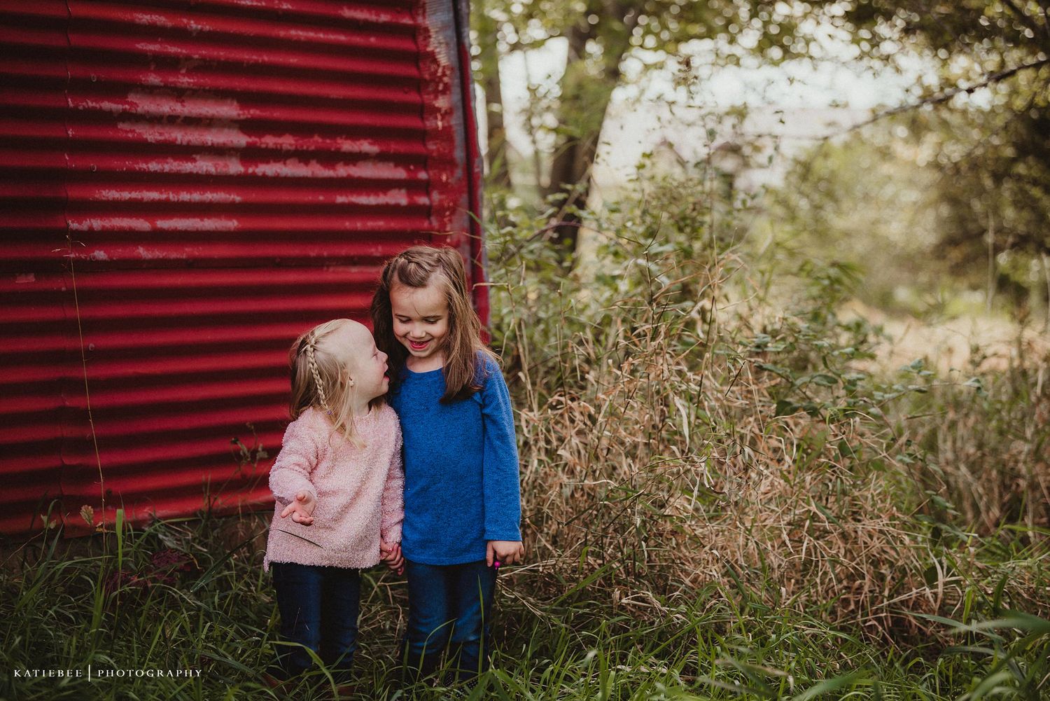 Two children in sweaters standing close together by a red barn wall.