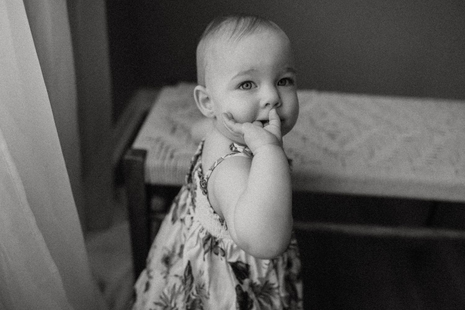 A black and white portrait of a small child in a patterned dress near a window.