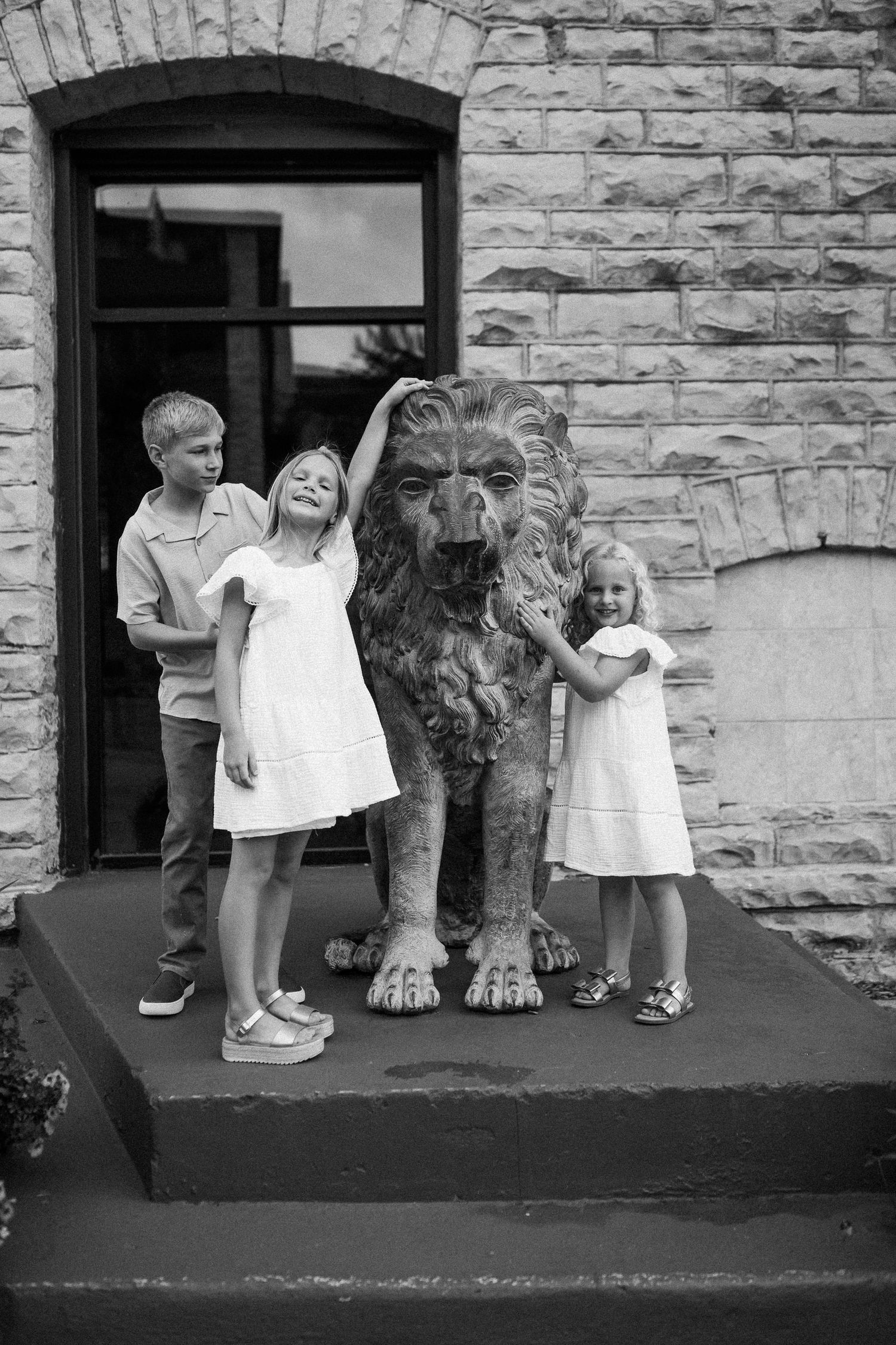 Children pose with a large stone lion statue at the entrance of a brick building in black and white.