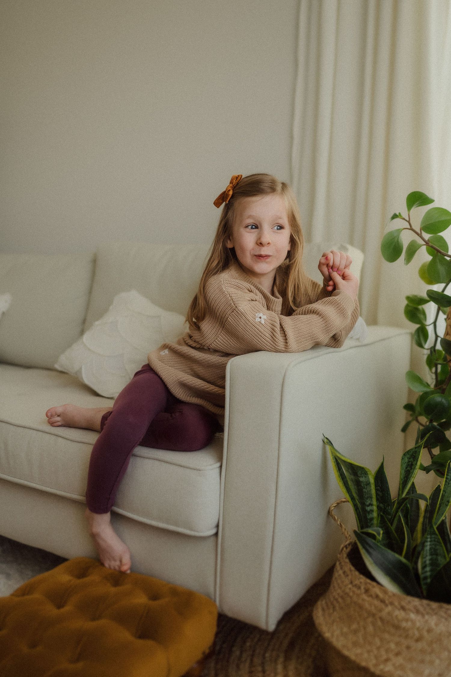 Candid lifestyle photo of child relaxing on white couch wearing beige sweater and burgundy leggings.