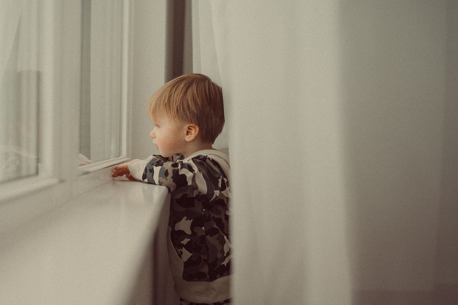 Child in camo print outfit peering out window in moody portrait.