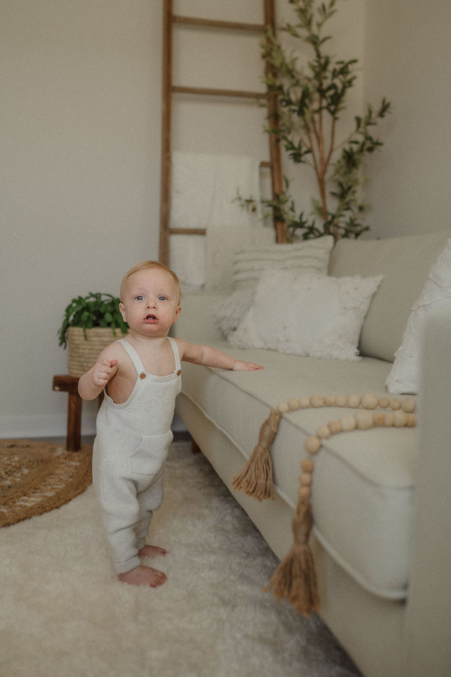 A small child in white overalls stands by a beige couch with decorative beaded garland.