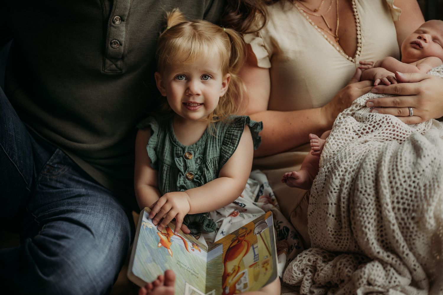 Toddler holds colorful book while sitting with family on floor.