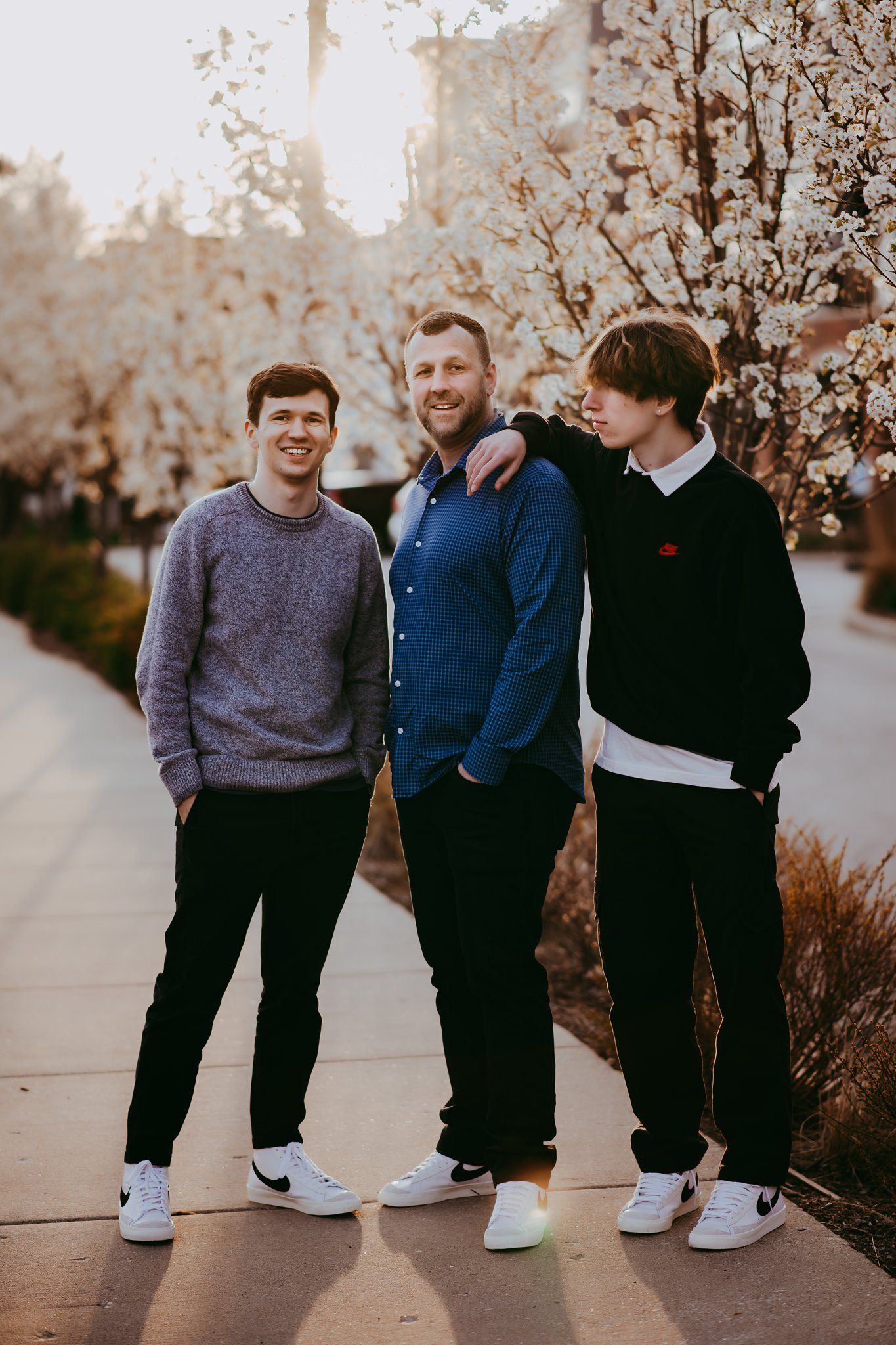 Three people smile together outdoors with blooming cherry trees in background during sunset.