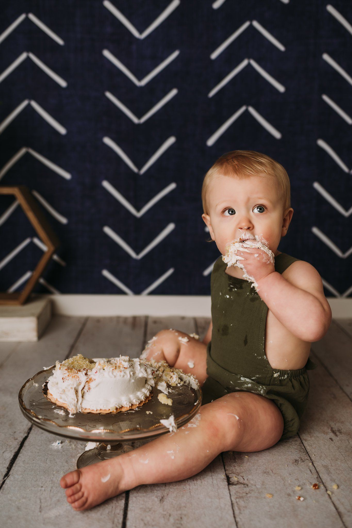 Child wearing olive romper sits on floor with smashed cake against navy chevron wall backdrop.