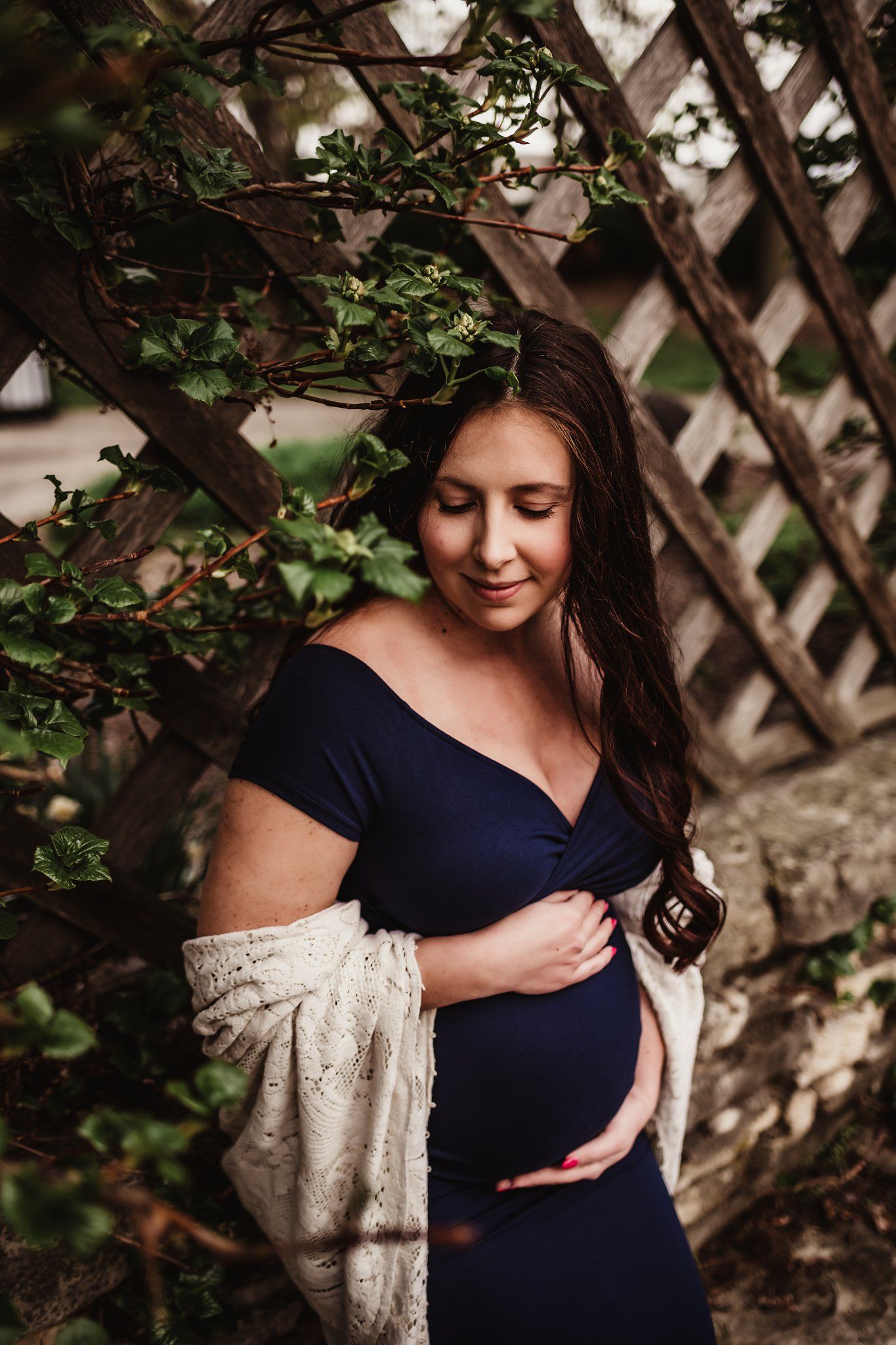 Maternity portrait with navy dress and cream cardigan against wooden trellis with vines.