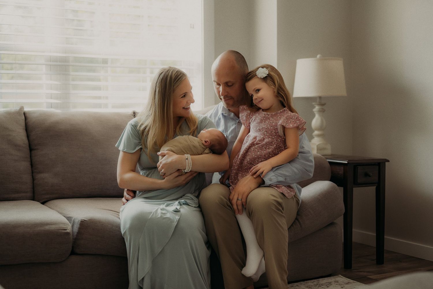 Family sitting together on grey couch while parent holds newborn baby.