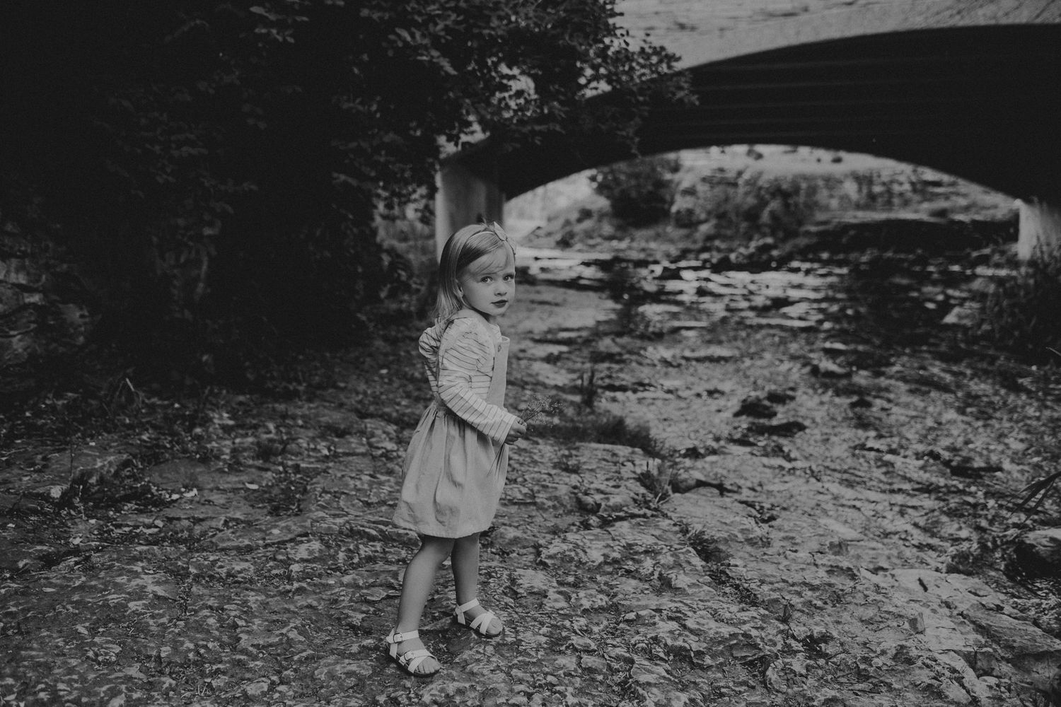 Black and white photo of a child standing beneath a stone bridge by a stream.