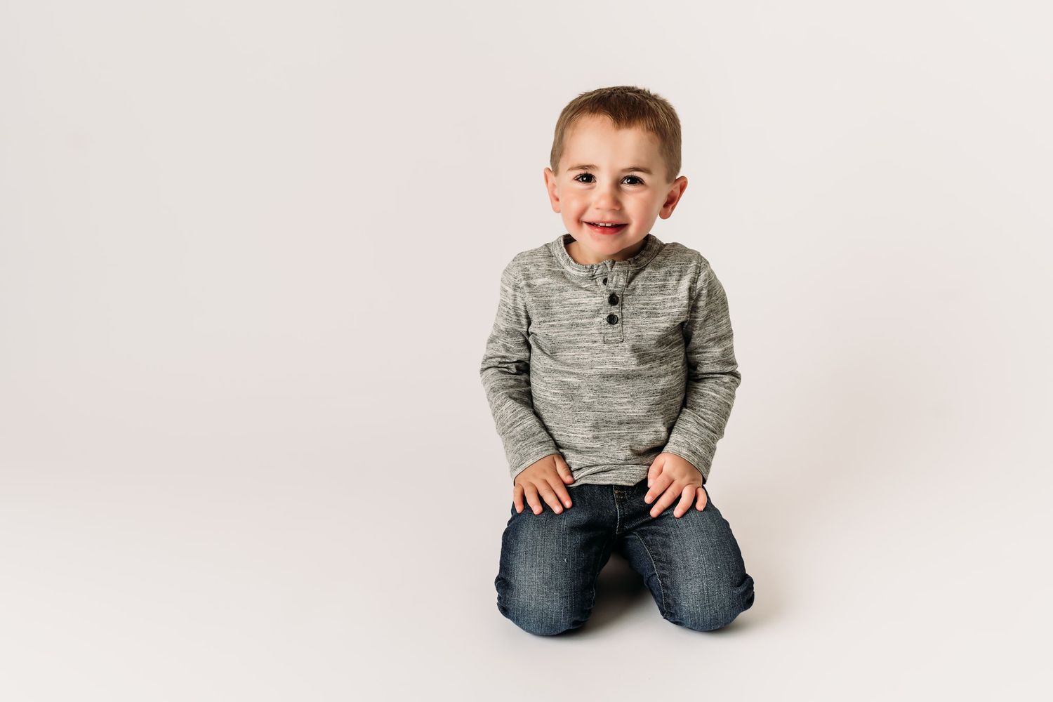 Cheerful kneeling pose on white background wearing gray henley shirt and jeans.