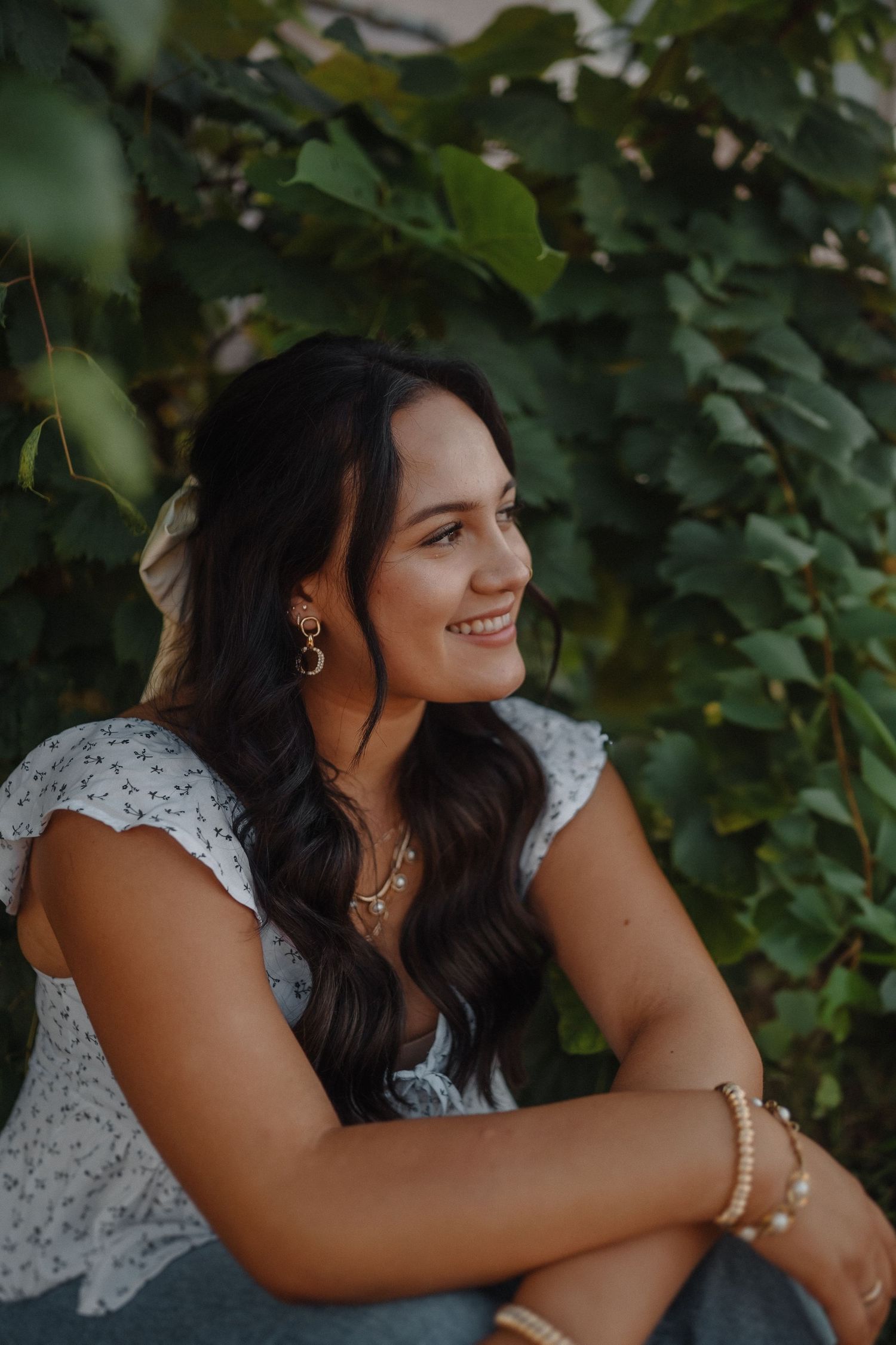 Portrait against green foliage showing someone in a white lace top with long dark wavy hair and jewelry.