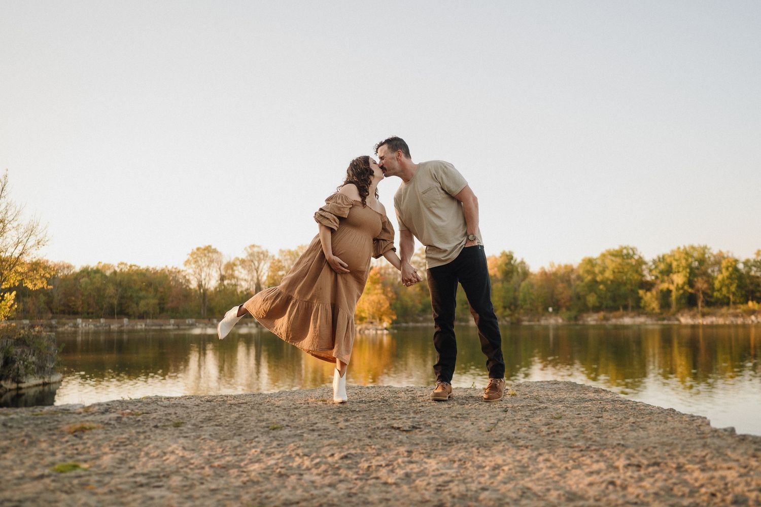 Romantic maternity photo of couple dancing by lake at golden hour.