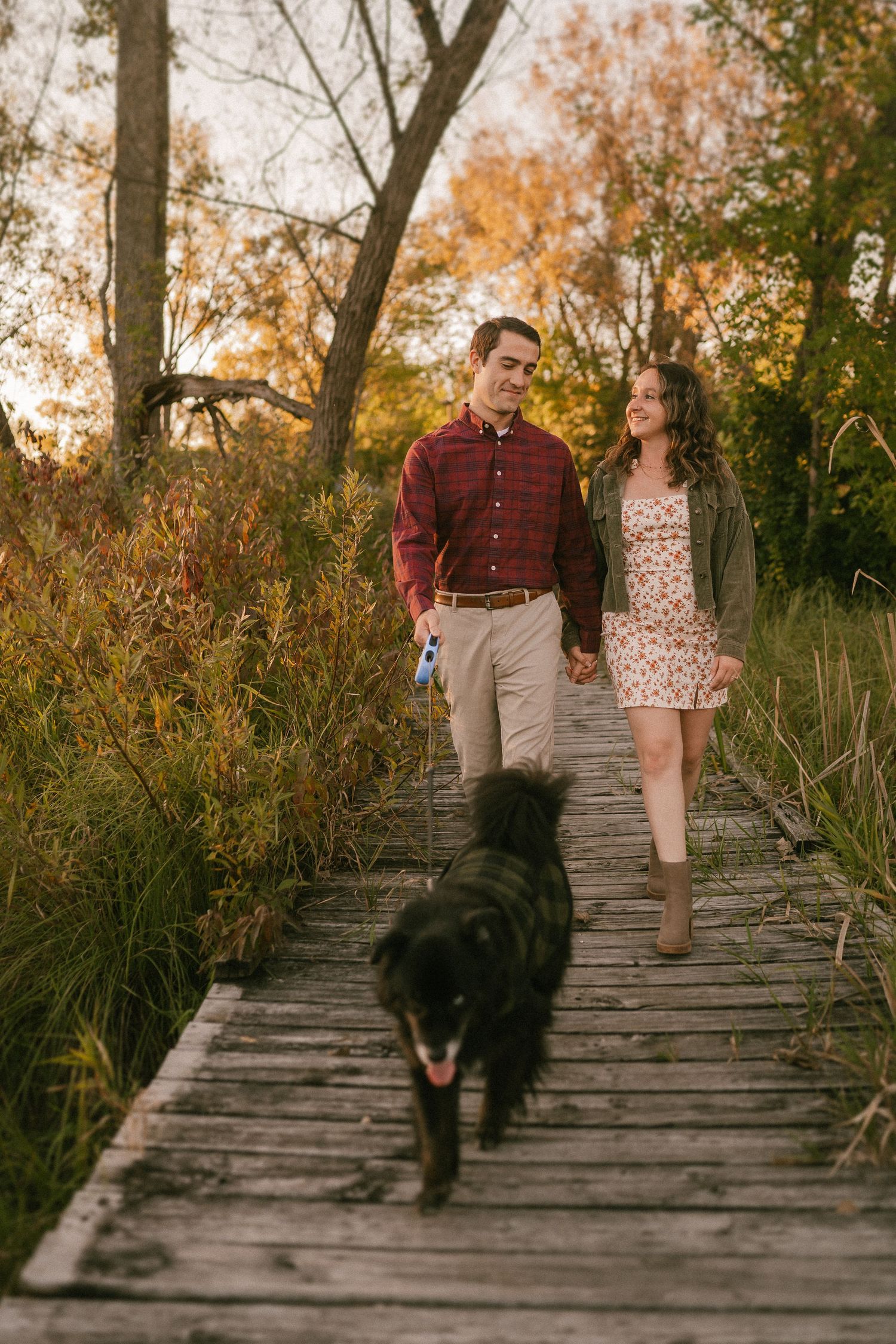 A couple walks their black dog along a wooden boardwalk path surrounded by autumn foliage.